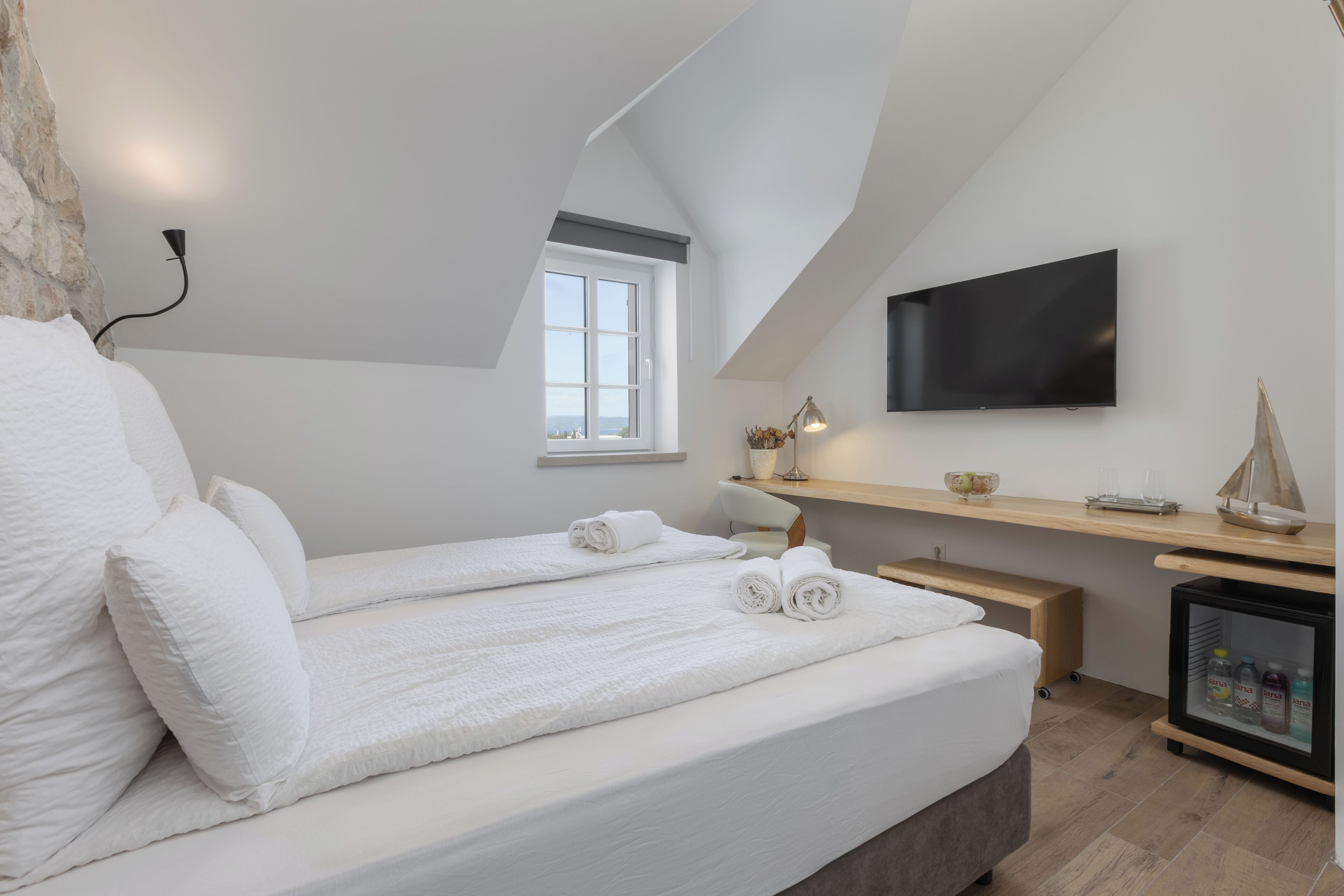 Une chambre à coucher moderne avec un grand lit, un téléviseur fixé au mur, une petite fenêtre et une étagère en bois sur laquelle sont posés divers objets.