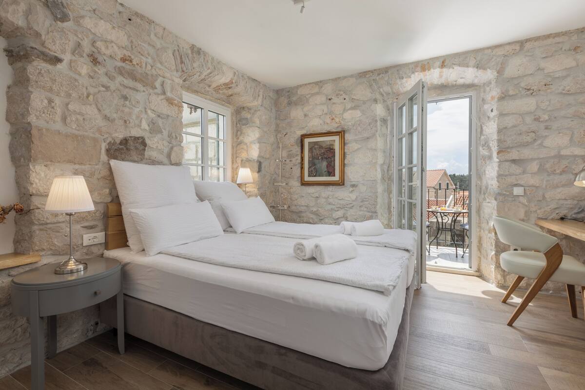 Ein Schlafzimmer mit Steinwand, einem Bett mit weißen Leinen, einem Nachttisch mit Lampe und Blick ins Freie.