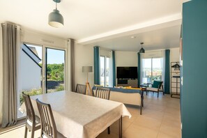 A modern dining area with a table and chairs, a TV, and a view of greenery outside.