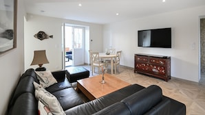 A modern living room with a black leather sofa, a wooden coffee table, and a TV mounted on the wall. There is a dining area with a table and chairs, and a wooden cabinet with drawers. - La Maison des Ours - proche plage de Courseulles (Courseulles-sur-Mer)