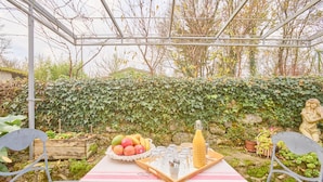 An outdoor dining setup with a table covered by a Paris-themed tablecloth, surrounded by greenery and a statue. - Pierre Idyllique - Climatisée avec grand jardin (Saumont)