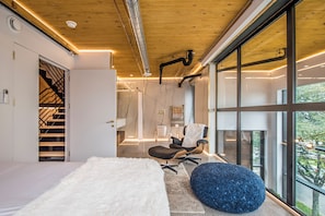 A modern bedroom with a large window, a white fluffy rug, a blue ottoman, and a chair.