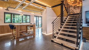 A spacious interior with a wooden staircase, a kitchen area with a bar, and large windows offering a view of greenery.