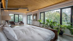 A bedroom with a large bed, two chairs, a potted plant, and a view of trees through large windows.