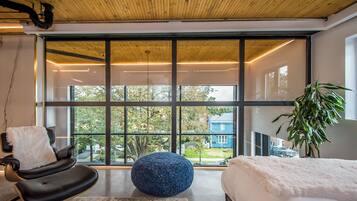A modern room with a large window, a black chair, a blue ottoman, and a bed with a white blanket.