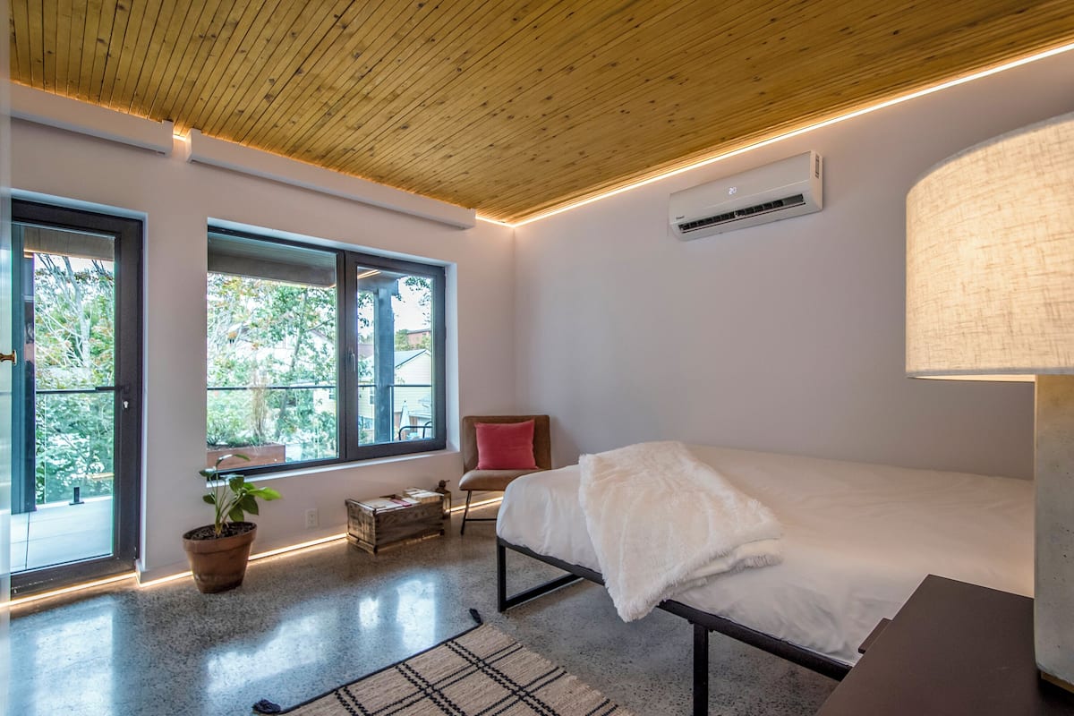 Un dormitorio moderno con techo de madera, ventanas amplias, una cama, una silla y una planta en maceta.
