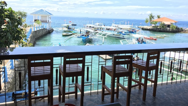 A terrace with wooden chairs and tables overlooking a harbor with boats and a small building. - CNC DIVE RESORT (Lapu-Lapu)