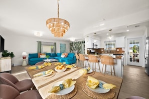Ein modernes Wohnzimmer mit einem blauen Sofa, einer Essecke mit einem Holztisch, der für eine Mahlzeit gedeckt ist, und einer Küche mit weißen Schränken und einem Edelstahlkühlschrank.