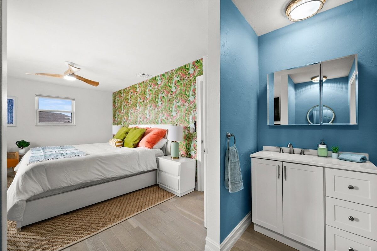 Ein Schlafzimmer mit Bett, Nachttischen, Deckenventilator, Fenster und einem Badezimmer mit Waschbecken, Spiegel und Handtuchhalter.