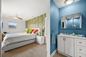 Ein Schlafzimmer mit Bett, Nachttischen, Deckenventilator, Fenster und einem Badezimmer mit Waschbecken, Spiegel und Handtuchhalter.