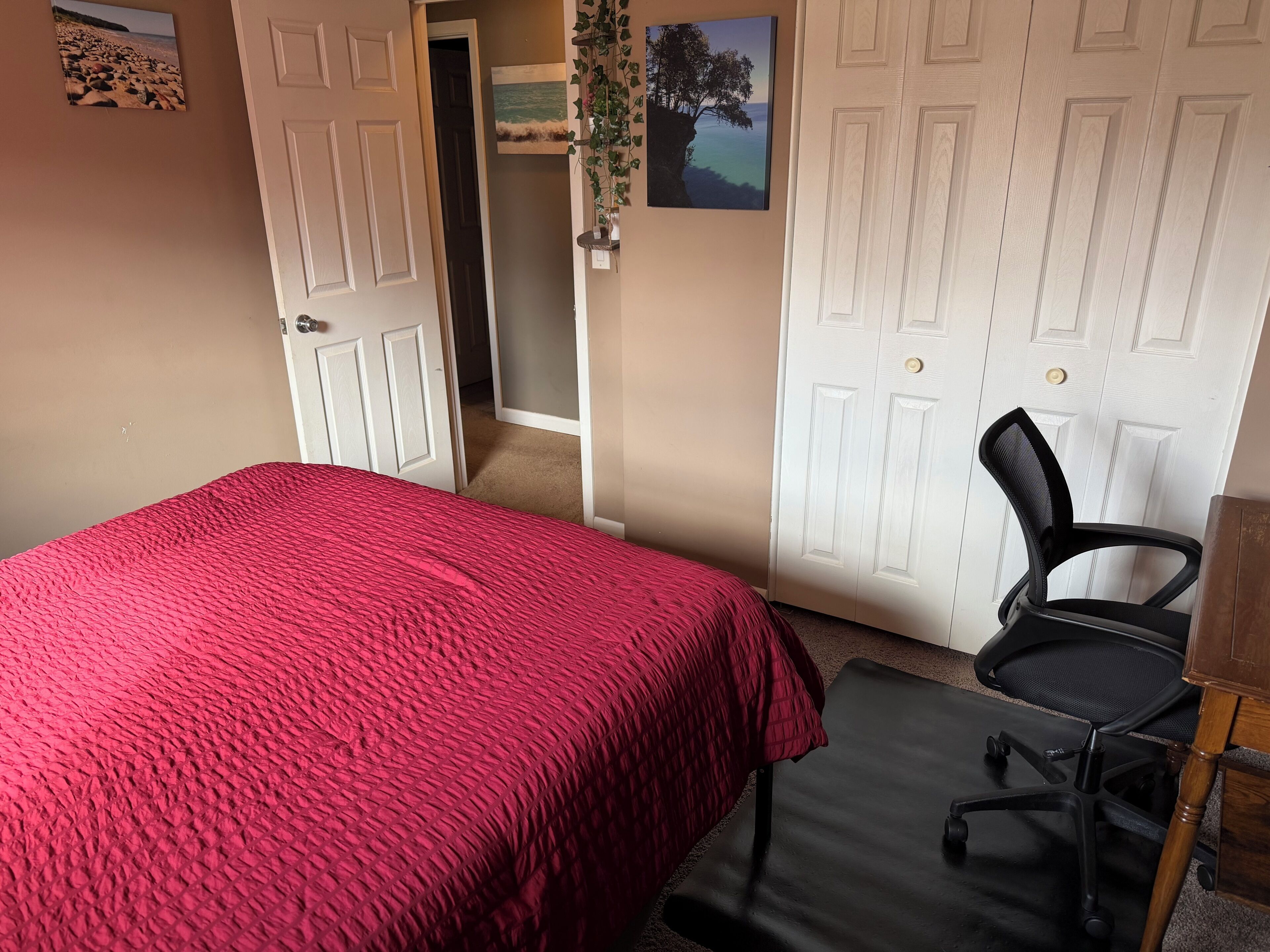 Une chambre à coucher avec une couvre-lit rouge, un fauteuil de bureau noir et une porte donnant sur une autre pièce.