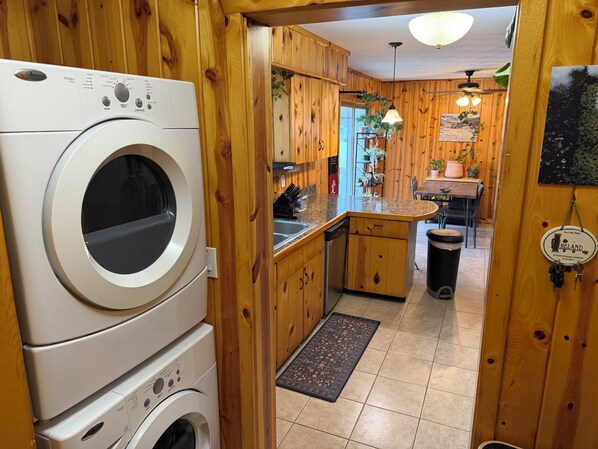 A laundry area with stacked washing machines, wooden cabinets, a kitchen area with a sink, and a trash bin. - Relax near CMU and Casino with hot tub and fireplace (Mount Pleasant)