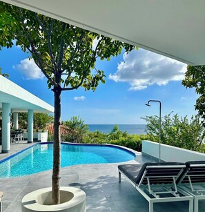 A modern pool area with a clear view of the ocean, a tree planter, and lounge chairs.