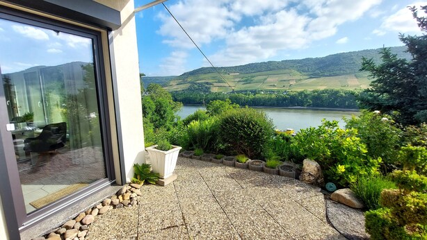 A modern building with a glass door, a gravel patio, and a view of a lake and hills. - Apartment Märchenhain with beautiful view (Niederheimbach)