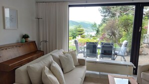 A living room with a sofa, a piano, and a view of a garden through a sliding glass door. - Apartment Märchenhain with beautiful view (Niederheimbach)