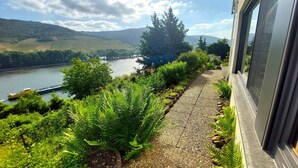 A view from a window overlooking a river, with a boat passing by and a path lined with greenery. - Apartment Märchenhain with beautiful view (Niederheimbach)