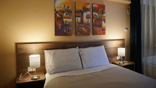 A hotel room with a wooden headboard, a bed with white linens, two bedside tables with lamps, and three paintings on the wall.