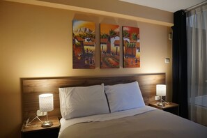 Een hotelkamer met een houten hoofdeinde, een bed met witte lakens, twee nachtkastjes met lampen en drie schilderijen aan de muur.
