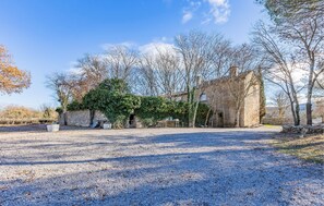 Un bâtiment historique en pierre, doté d’une cour en gravier, entouré d’arbres et surplombé d’un ciel dégagé.
