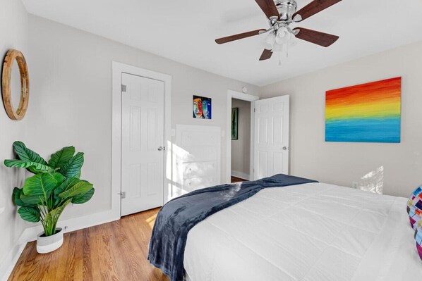Une chambre à coucher avec un lit, un ventilateur de plafond, une grande plante et un tableau coloré au mur.