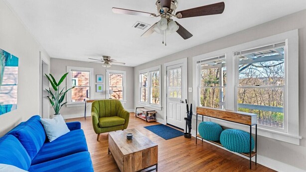 Een ruime woonkamer met een blauwe bank, een groene fauteuil, een houten salontafel en een plafondventilator. Grote ramen bieden natuurlijk licht en uitzicht op het buitenleven.