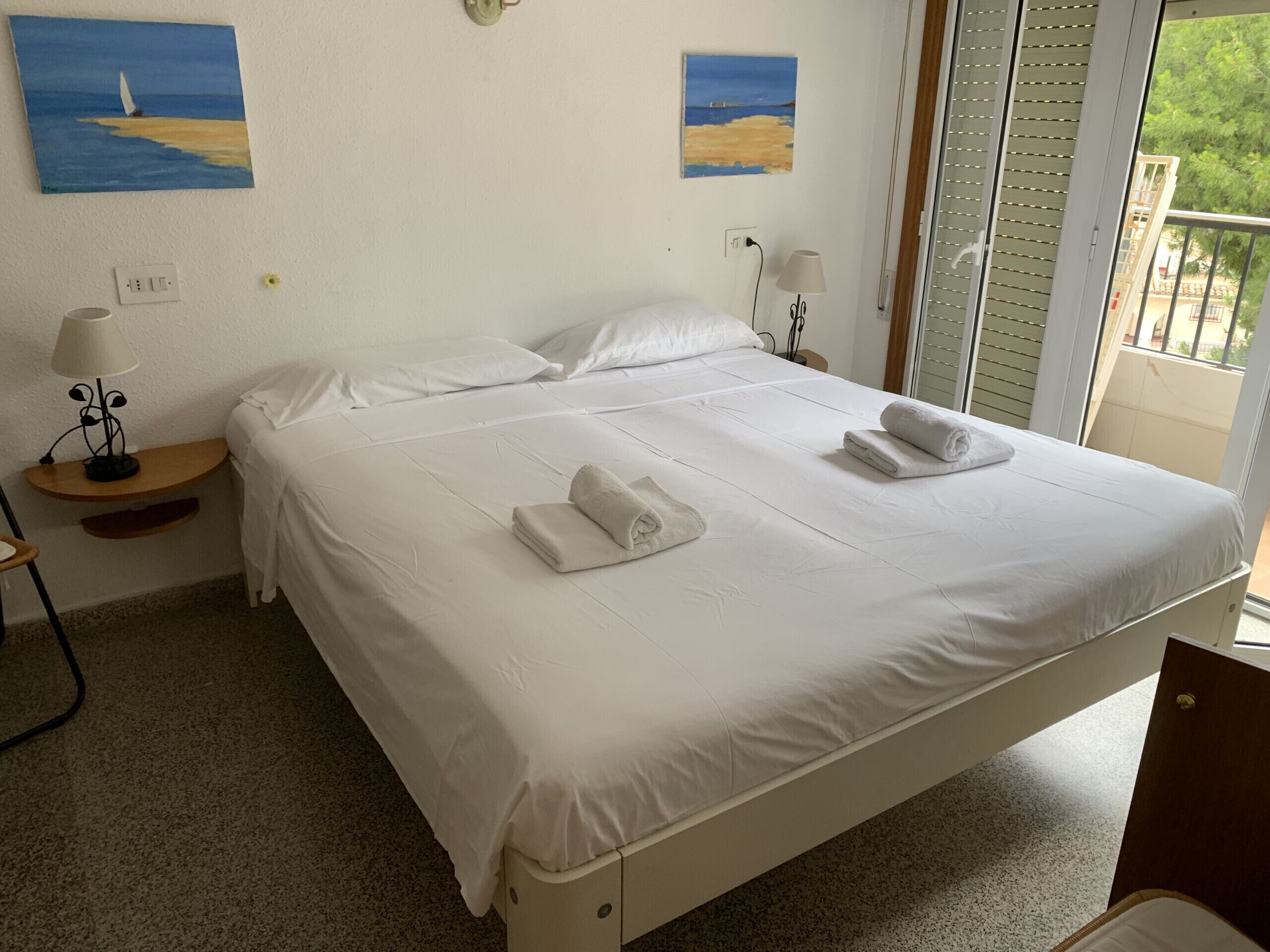 Quarto de hotel com cama, duas pinturas, uma luminária e janela com persianas.