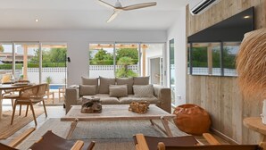 Ein modernes Wohnzimmer mit einer Couch, einem Couchtisch und Sesseln. An der Wand ist ein Fernseher angebracht und eine Deckenleuchte sorgt für Luftzirkulation.