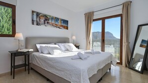 A bedroom with a large bed, two bedside tables, a painting on the wall, and a view of the mountains through the window. - Villa Vinalopo La Sella (Pedreguer)