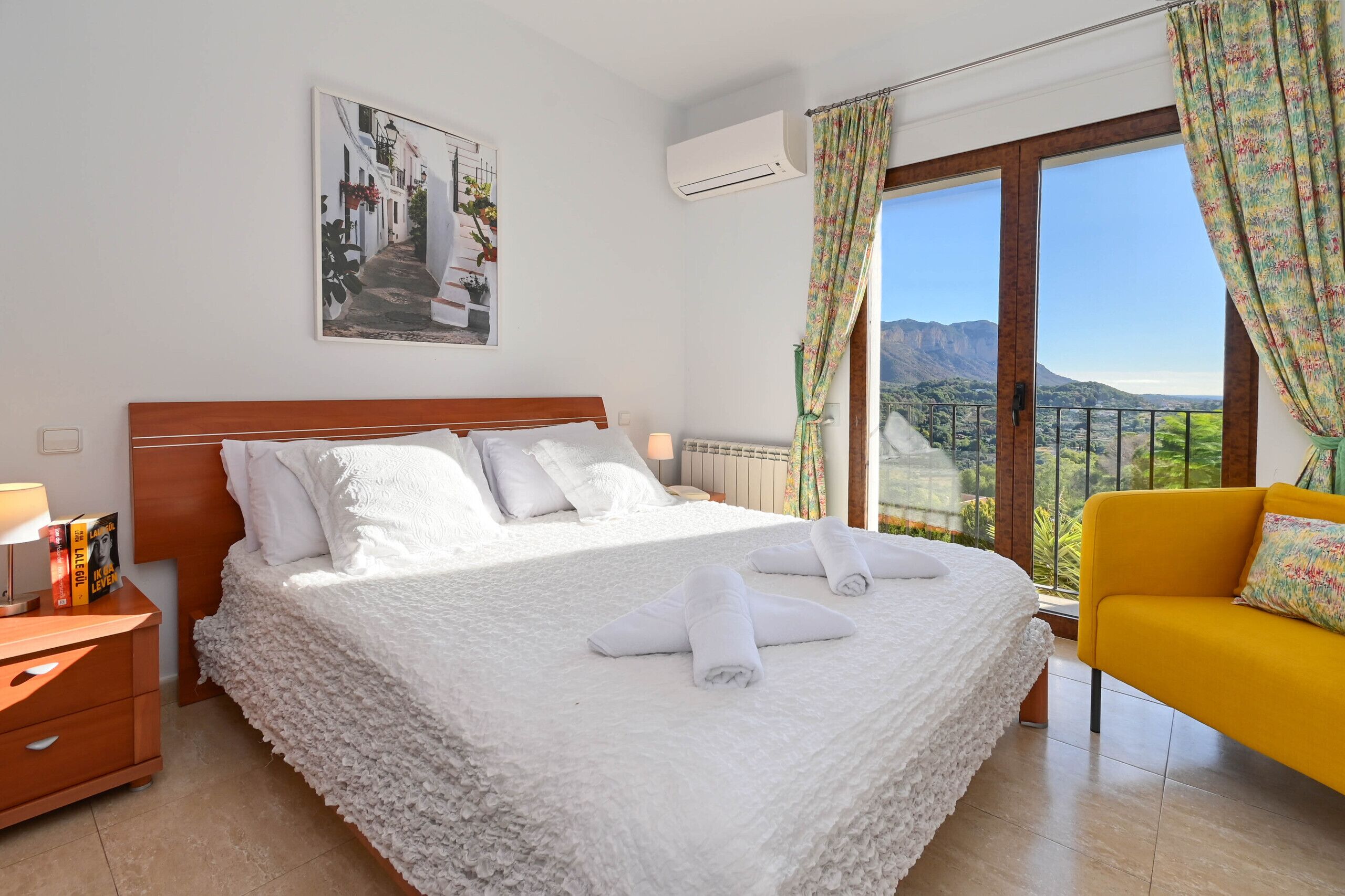 Ein Hotelzimmer mit einem großen Bett, einem hölzernen Nachttisch, einem gelben Sessel und Blick auf Berge durch das Fenster.