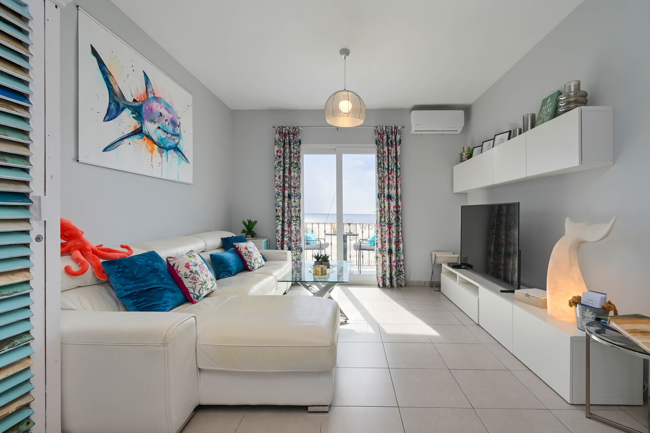 Uma sala de estar moderna com um sofá branco, uma mesa de centro de vidro e uma TV sobre um suporte branco. Há uma varanda com vista, cortinas e um lustre.