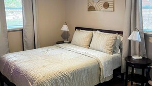 Una cama bien hecha con edredón blanco y almohadas, un ventilador de techo, una mesita de noche con una lámpara y un adorno en la pared.