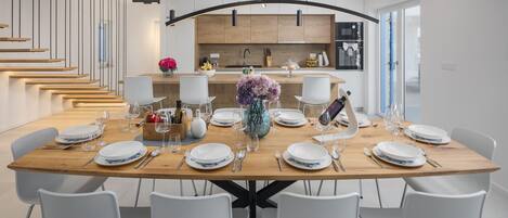 A modern dining area with a wooden table set for six, white chairs, and a kitchen in the background.