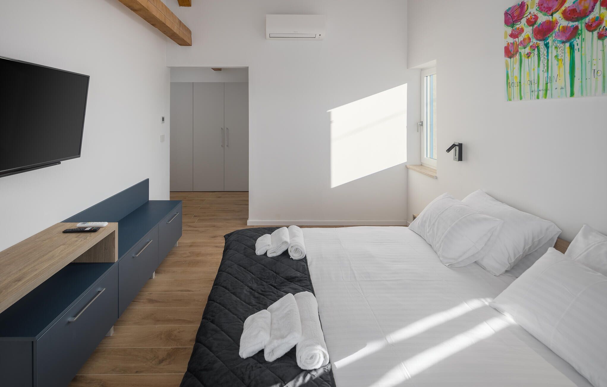 A modern hotel room with a bed, a TV, a desk, and a window with natural light.