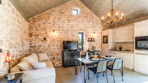 A cozy interior with a stone wall, a dining area with chairs and a table, a sofa, a TV, and a kitchen with cabinets and appliances.