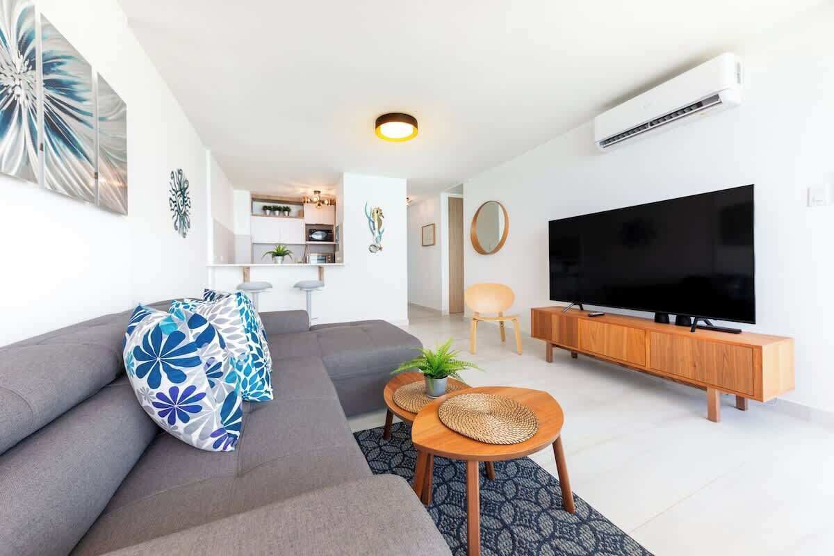 Una sala de estar moderna con un sofá gris, un televisor de pantalla plana y un mueble bajo para la televisión. Se ve una cocina al fondo con armarios blancos y una zona de comedor.