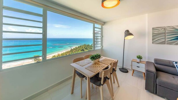 A modern living room with a large window offering a view of the ocean and beach. - Cozy BEACH FRONT 1BR w/Pool & PK @Luquillo Beach (Luquillo)