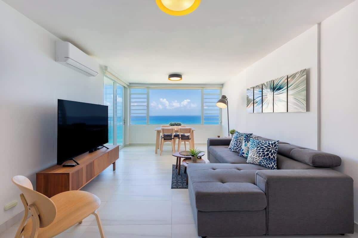A modern living room with a flat-screen TV, grey sofa, wooden chair, and a dining table with chairs.