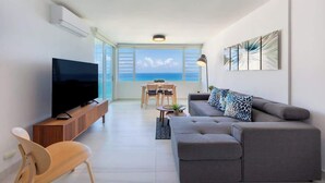 A modern living room with a flat-screen TV, grey sofa, wooden chair, and a dining table with chairs. - Cozy BEACH FRONT 1BR w/Pool & PK @Luquillo Beach (Luquillo)