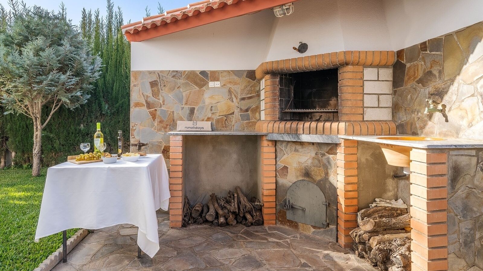 Eine Außenküche mit Steinwand, Backofen aus Ziegeln und Tisch, gedeckt mit Speisen und Getränken.