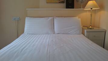 A neatly made bed with white linens, a bedside table with a lamp, and a wall-mounted power outlet.