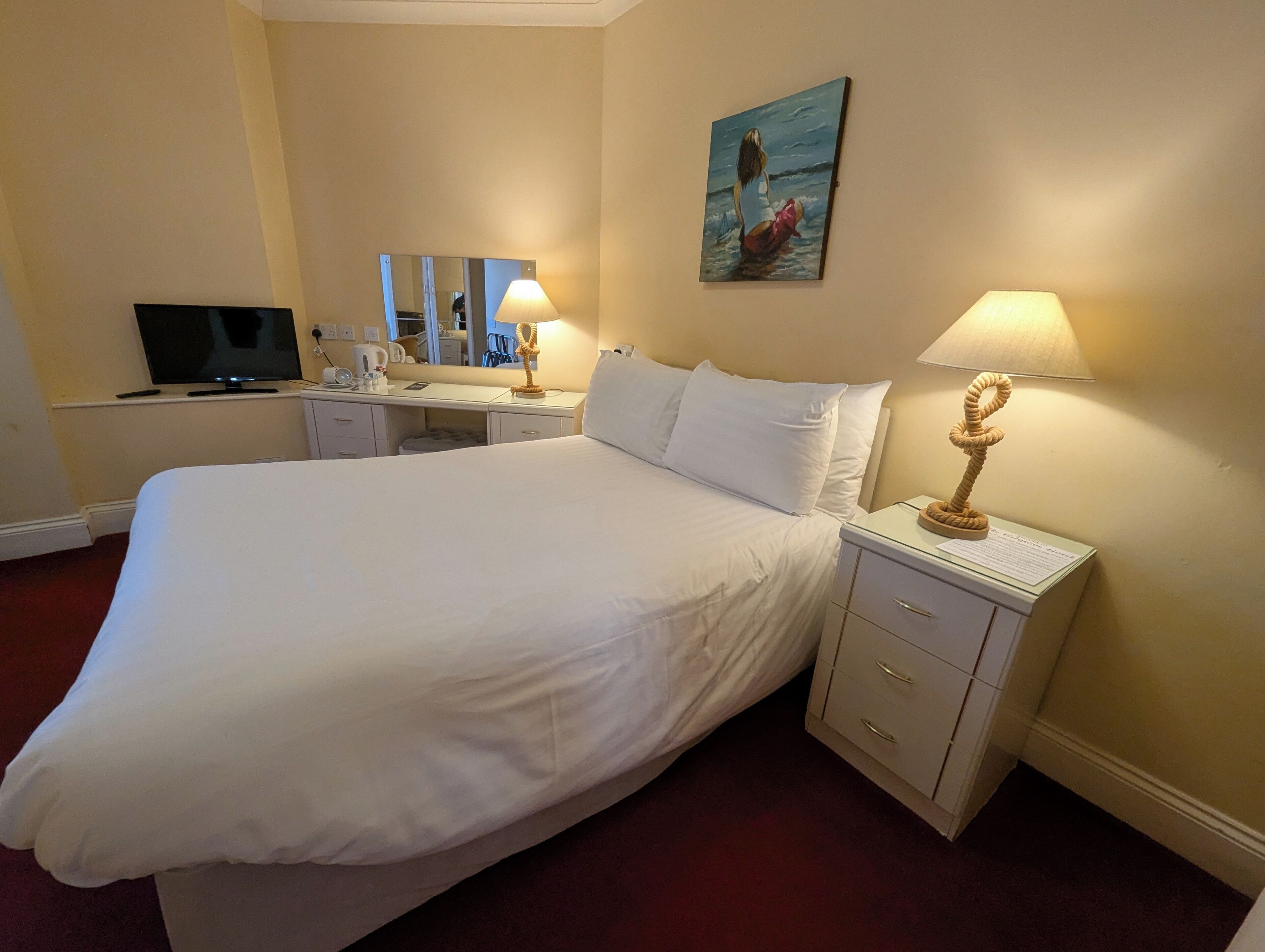 A hotel room with a large bed, a desk with a computer, a lamp, and a painting on the wall.
