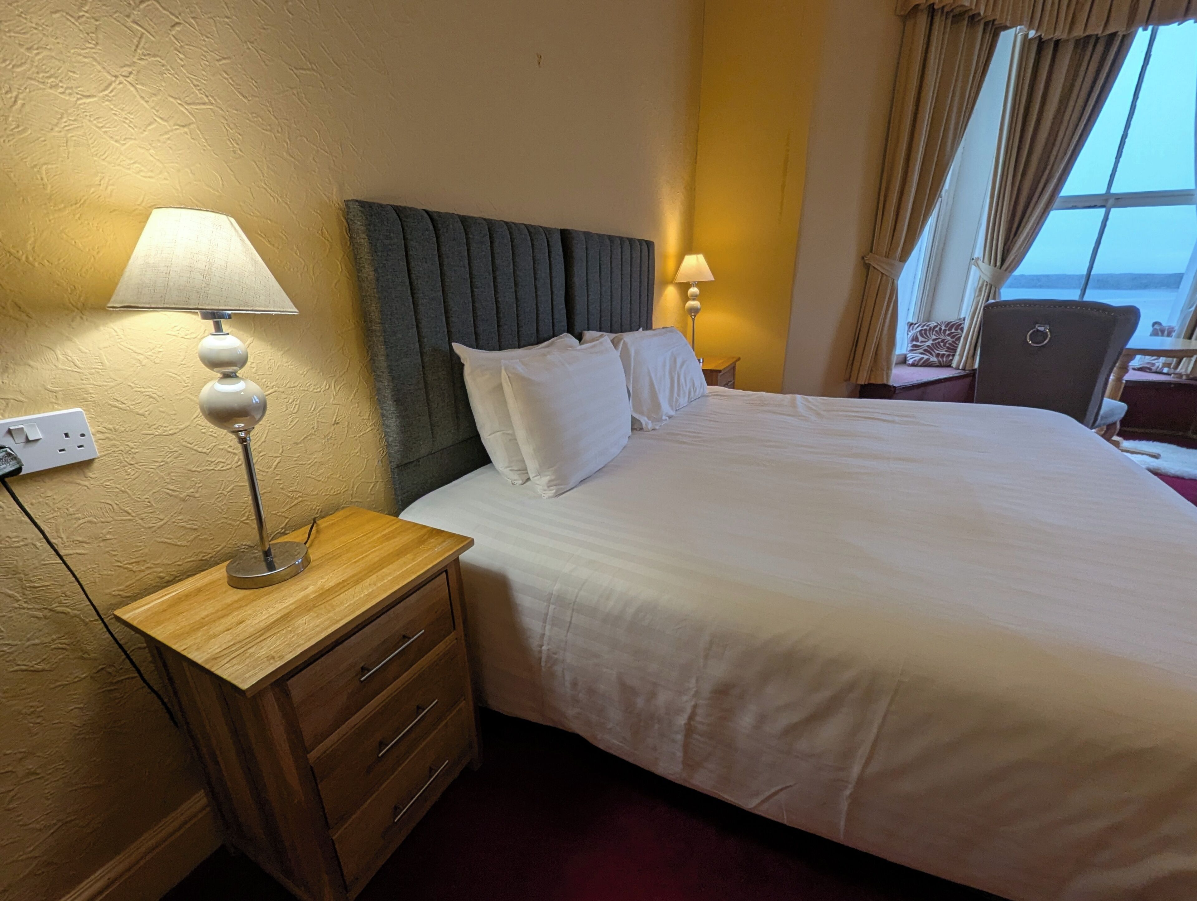 A hotel room with a large bed, two bedside lamps, a wooden nightstand, and a view of the sea through a window with curtains.