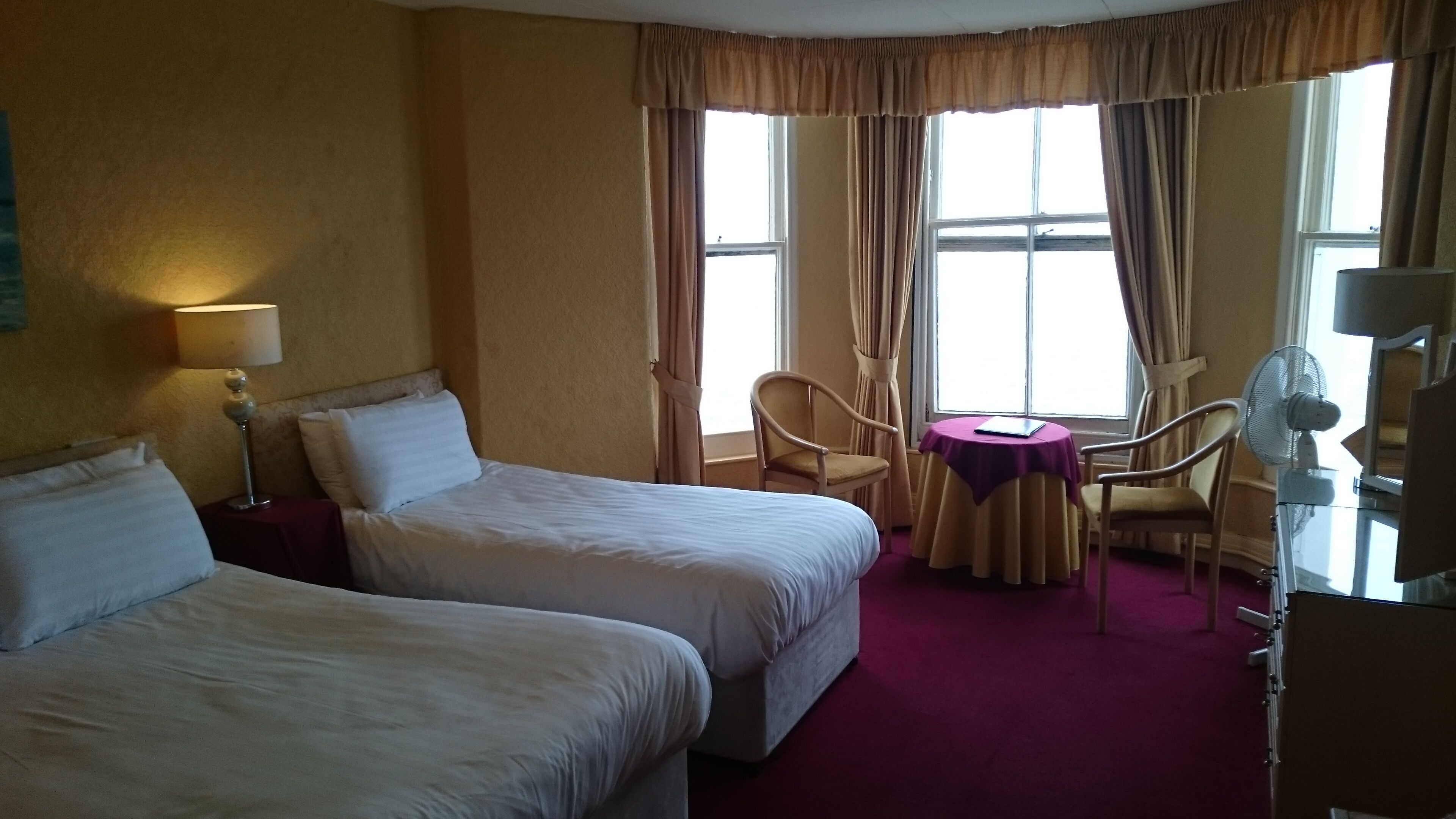 Quarto de hotel com duas camas, uma mesinha com uma cadeira, uma escrivaninha com cadeira e uma janela com cortinas.