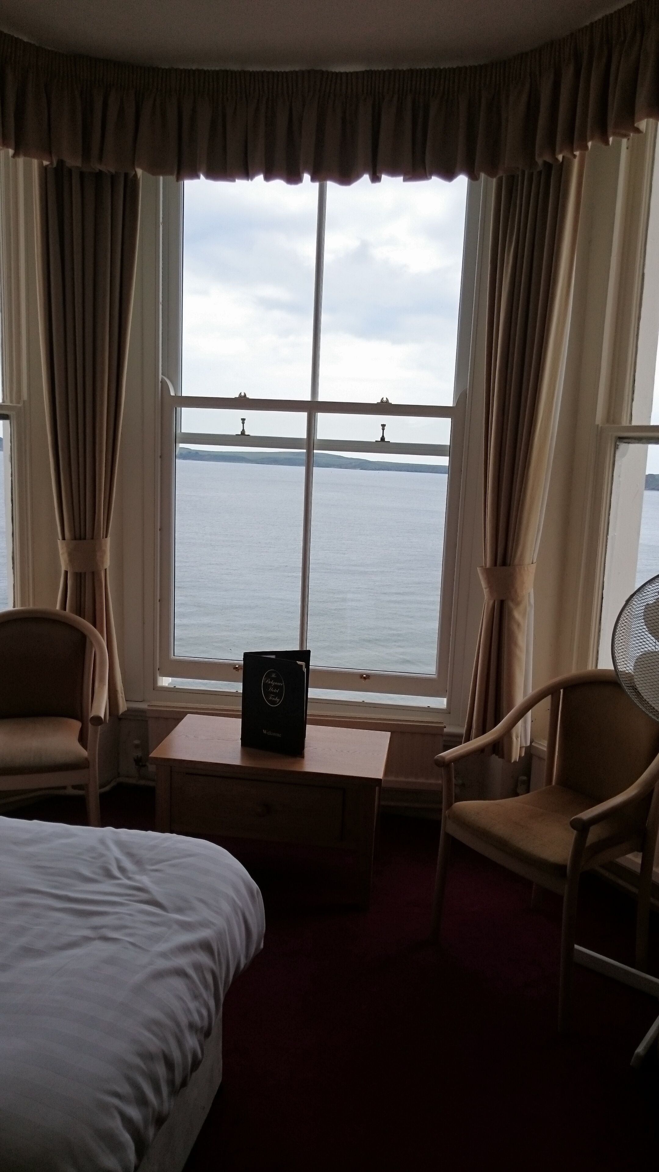 Une chambre d’hôtel avec un lit, deux chaises, une petite table sur laquelle est posé un panneau d’accueil, et une vue sur la mer grâce à de grandes fenêtres.