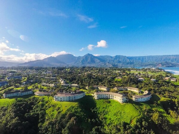 A resort with multiple white buildings, lush green surroundings, and a backdrop of mountains. - Alii Kai II 10A (Princeville)