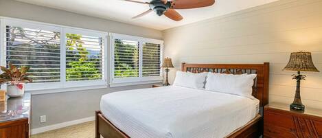 Un dormitorio con una cama de madera, un ventilador de techo, dos mesitas de noche con lámparas y una ventana con persianas.