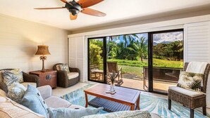 Un salón amplio con ventilador de techo, muebles de mimbre, una mesa de centro y vistas a un jardín exuberante a través de puertas corredizas de vidrio.