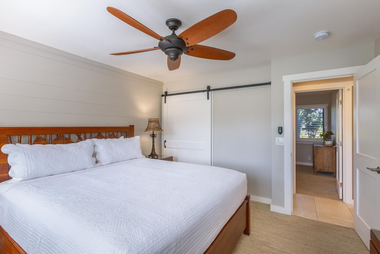 Un dormitorio con cama, ventilador de techo, mesita de noche, lámpara, puerta corredera y vista a un balcón.