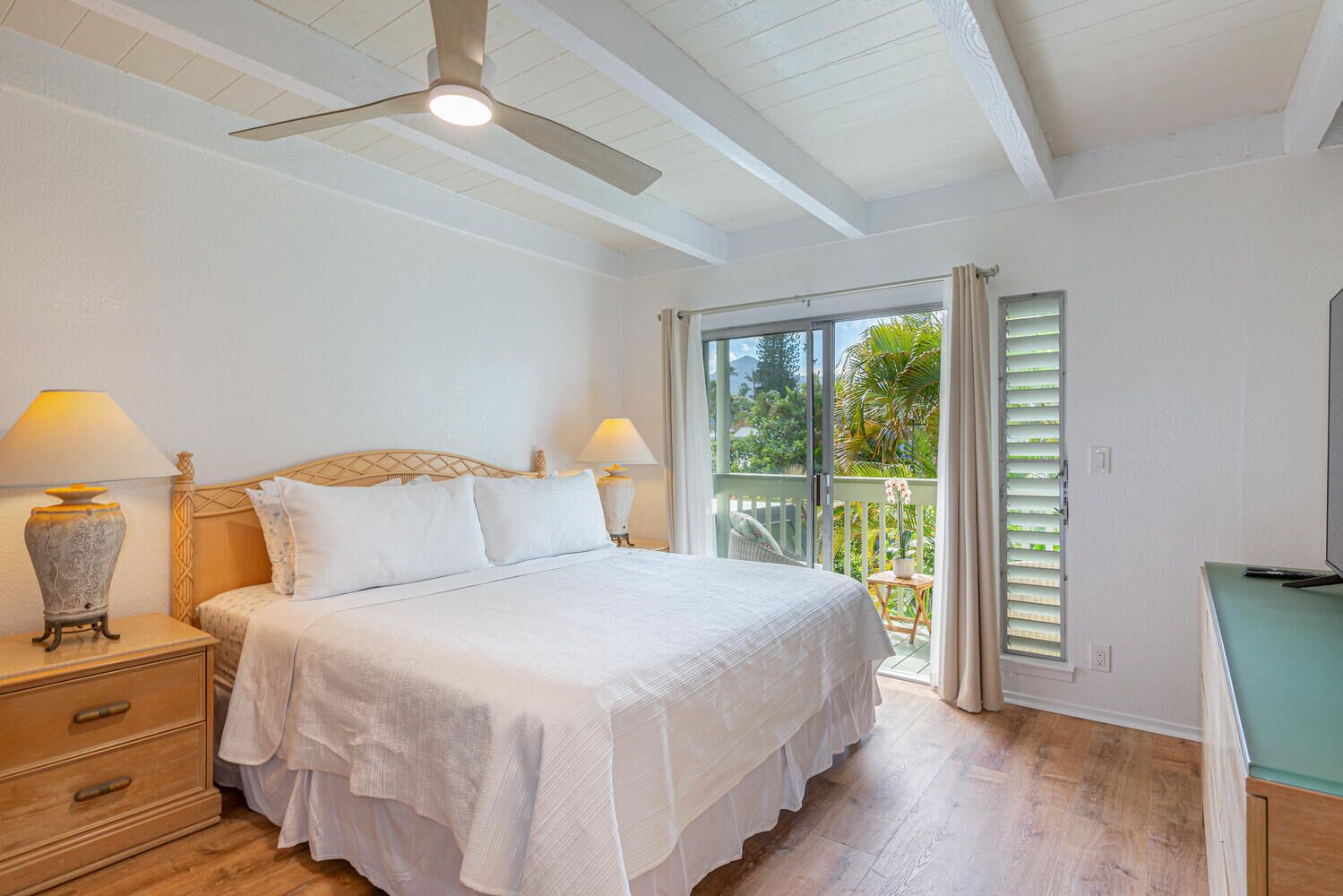 A bedroom with a bed, bedside tables, a ceiling fan, and a balcony with a view of greenery.