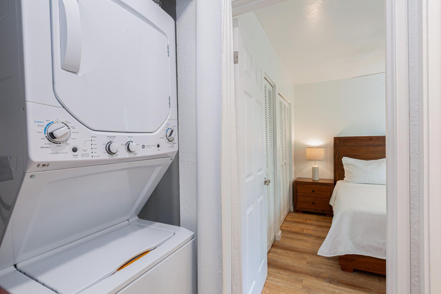 A hotel room with a washer and dryer, a wooden nightstand, a lamp, and a bed with white linens.